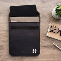 Black security and privacy pouch for tablets and iPads with multiple compartments, placed on a wooden desk with accessories.